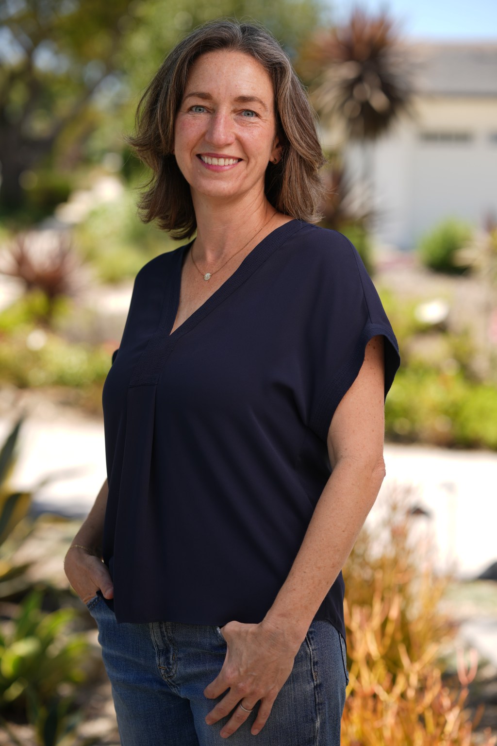 photo of white woman around 50 standing outside, brown hair and blue eyes, blue top and jeans with hands in pockets